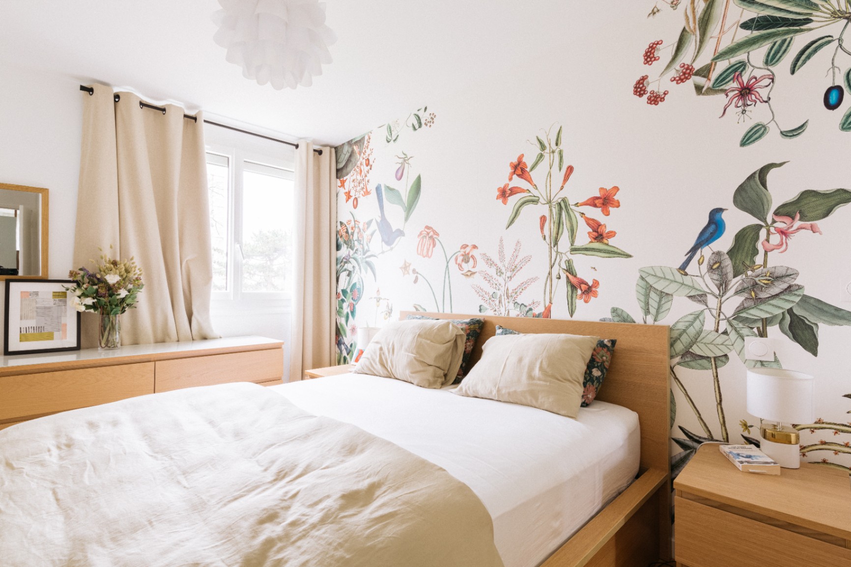 Chambre lumineuse avec lit en bois clair, linge beige et mur décoré d’un papier peint botanique ; commode avec vase de fleurs à gauche et table de chevet avec lampe à droite.