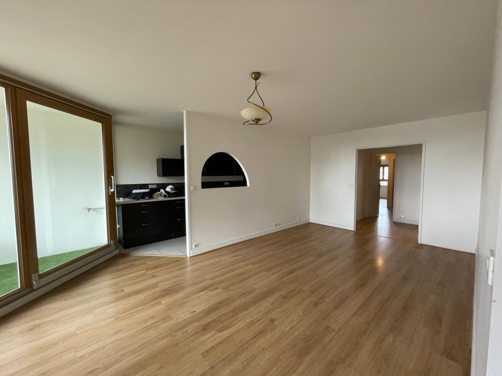 Séjour vide et lumineux avec parquet en bois clair, murs blancs, grande baie vitrée donnant sur un balcon, ouverture arrondie vers la cuisine et circulation fluide entre les espaces.