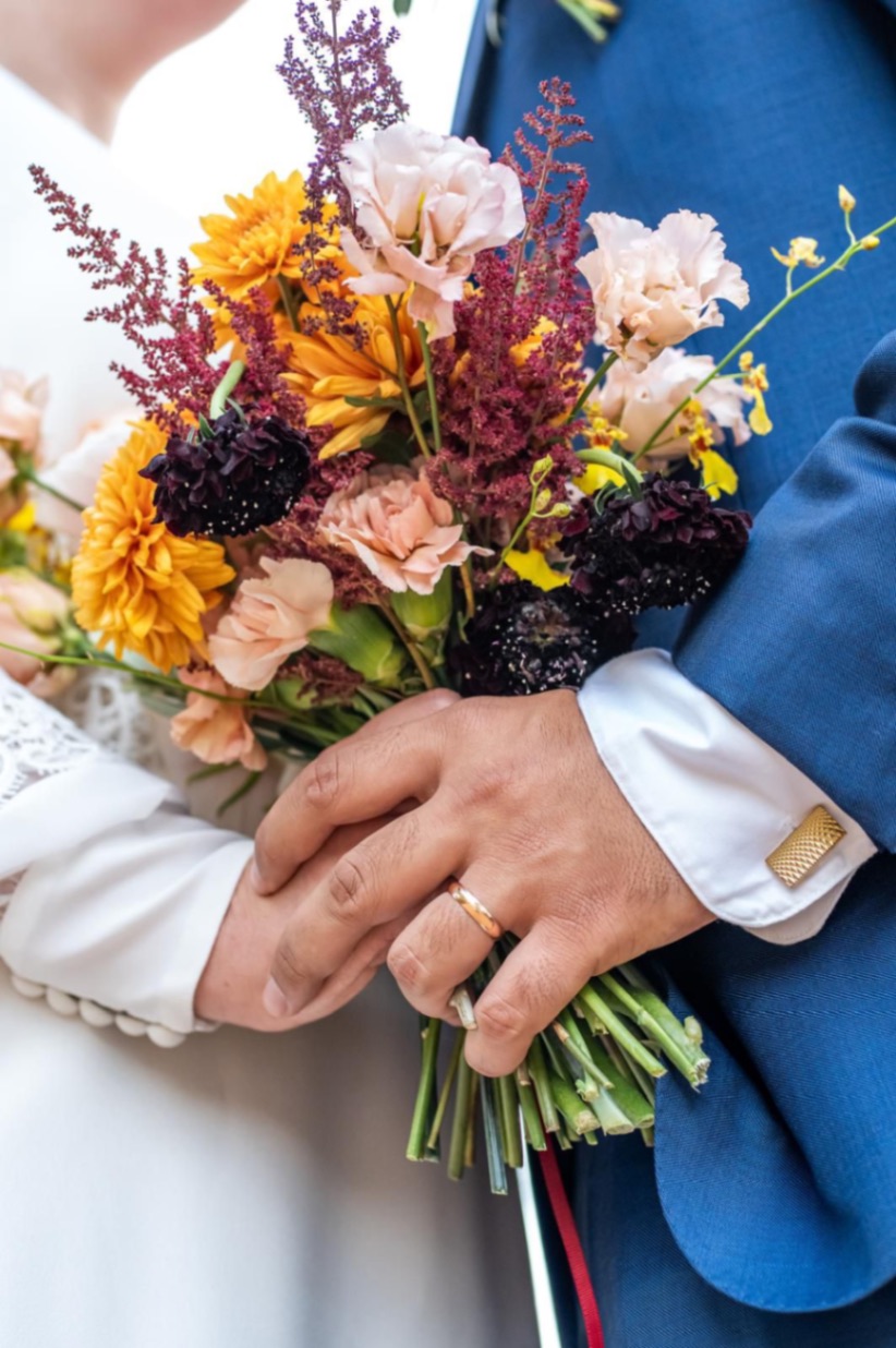 Bouquet de mariée pris en photo avec les alliances dans les mains des mariés, dahlia orangé, astilbe framboise, bouquet automnale avec scabieuse bordeau.jpeg